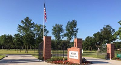 Entrance to Allen Memorial Park with gate, brick pillars, American flag, and sign under a blue sky.