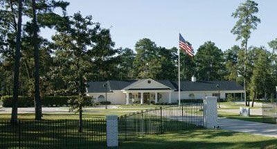 A white building with a US flag, trees, and a wrought-iron fence on a green lawn.