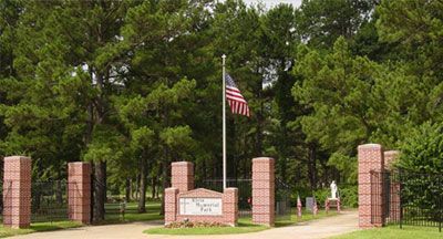 Entrance to a park with brick pillars, a sign, American flag, and trees.