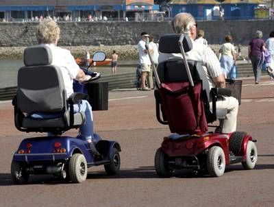 Two people are riding mobility scooters on a street.