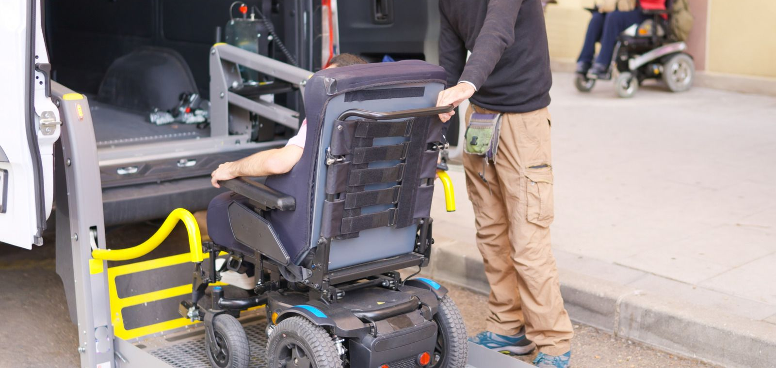 Person in a wheelchair being loaded into a vehicle, with another person assisting at the curb.