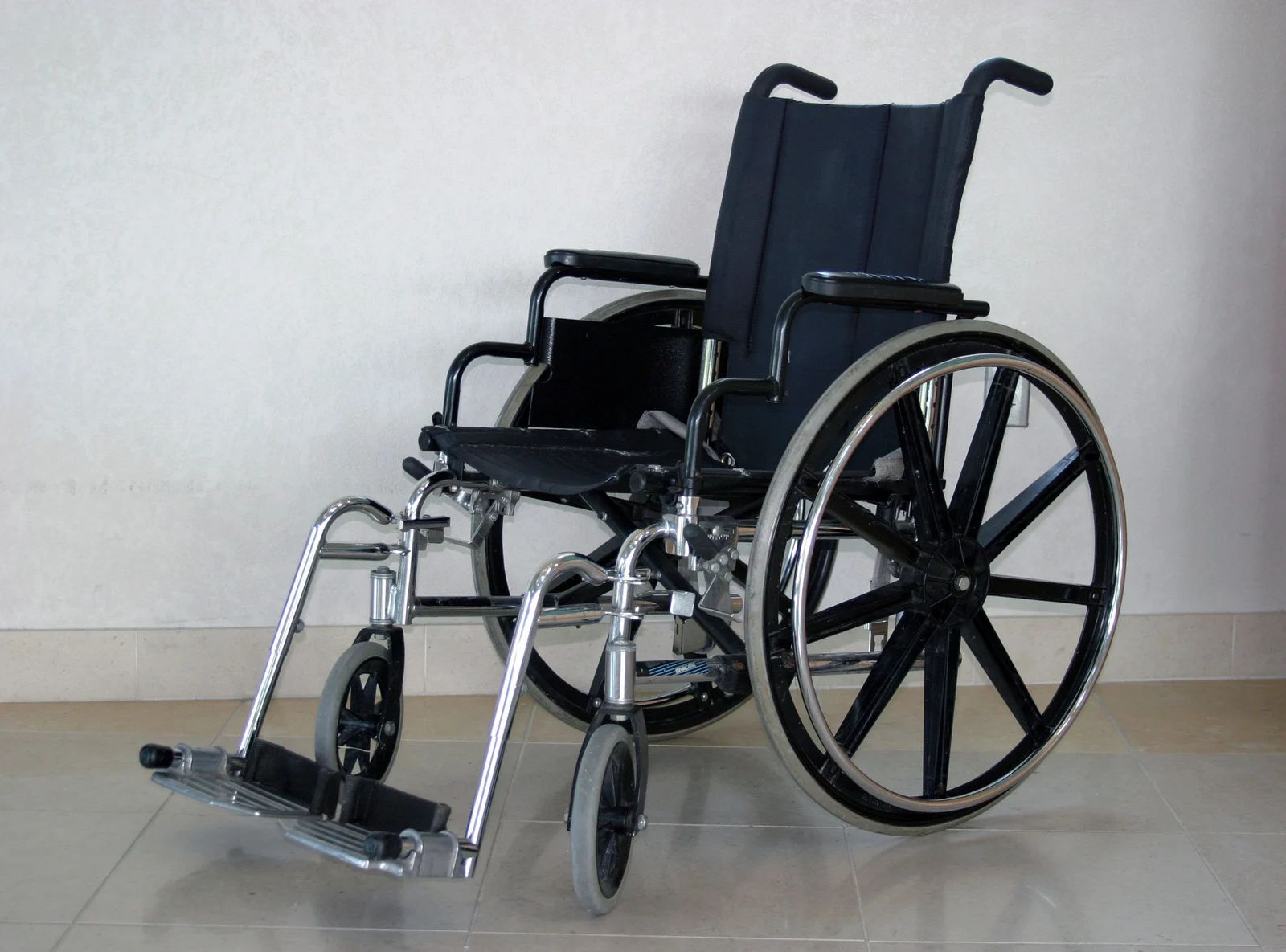 Black wheelchair with large rear wheels and front footrest against a plain wall.
