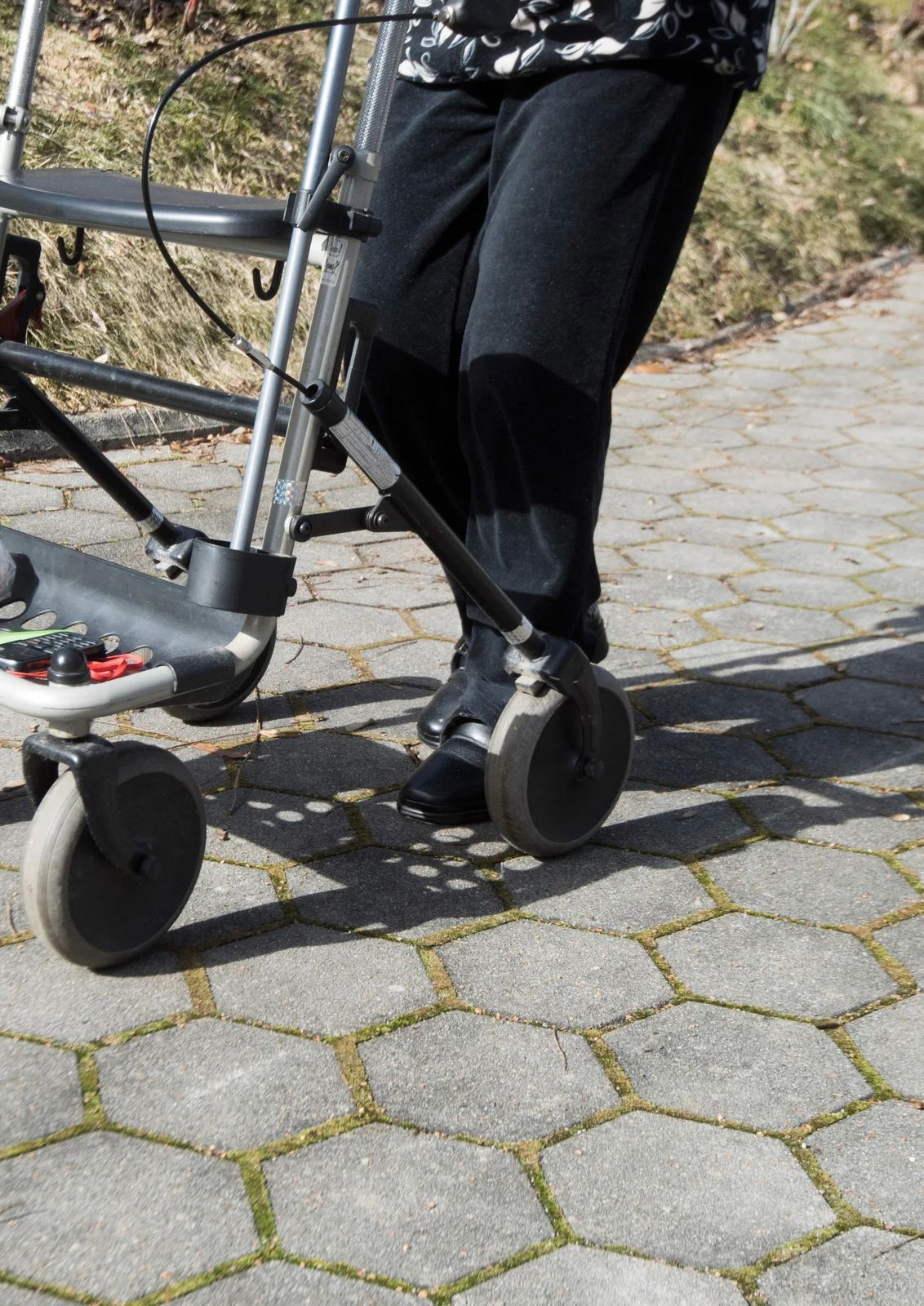 Person using a walker on a paved surface. Black pants, shadows visible.