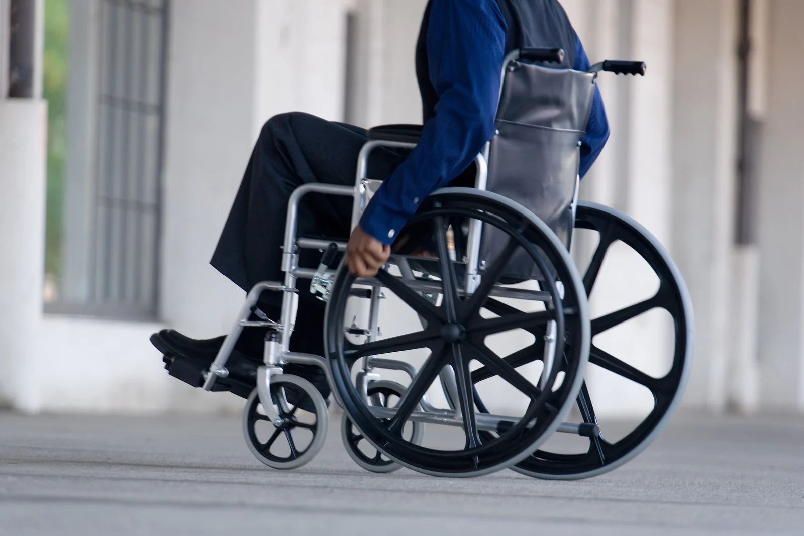 Person in a wheelchair indoors, wearing a blue jacket and black pants.