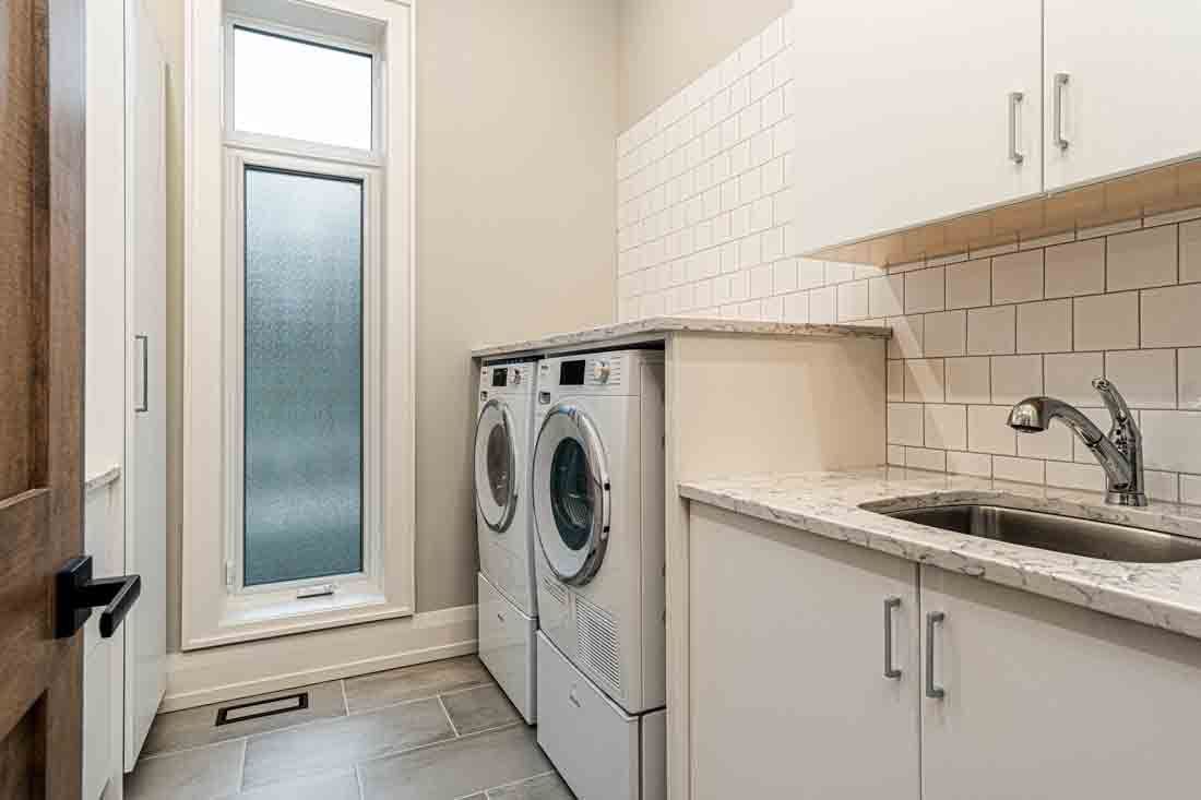 Bright laundry room with front-loading washer and dryer, white cabinets, sink, and frosted glass door.