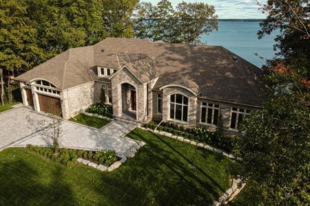Aerial view of a stone house with a driveway, lawn, and water behind it