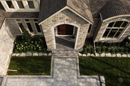 Front entrance of a stone house with arched doorway, paved walkway, and manicured lawn and shrubs