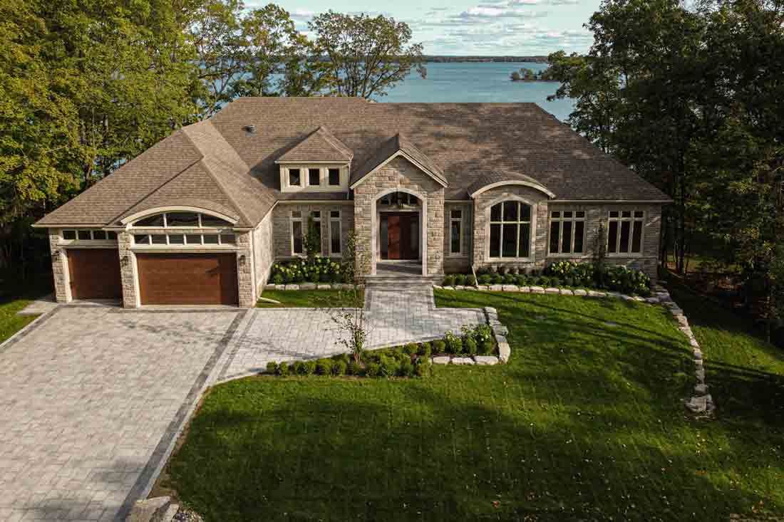 Aerial view of a large stone house with a driveway, green lawn, and water visible behind it.