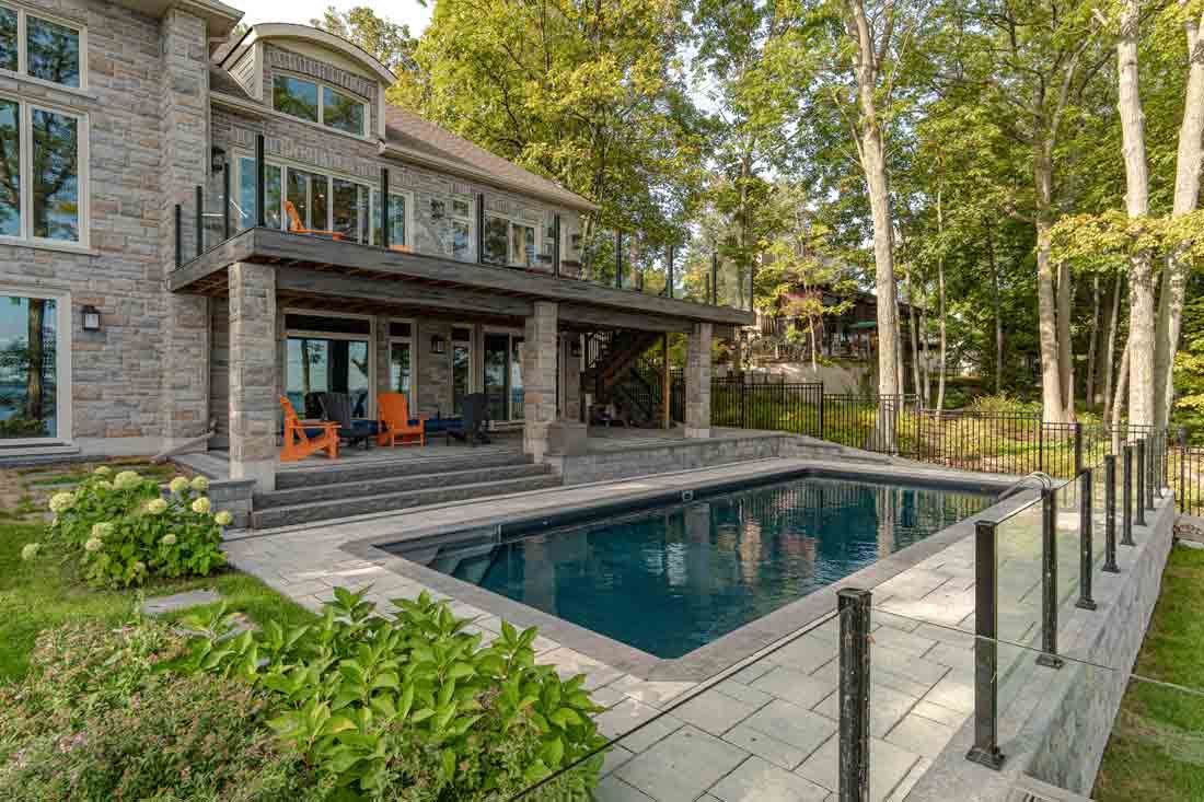 Stone house with a covered patio and rectangular backyard pool surrounded by trees and a glass railing