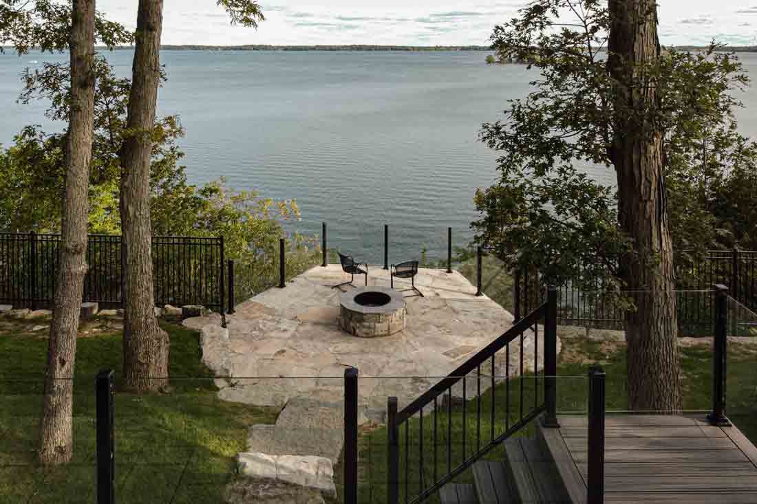 Stone fire pit overlooking a calm lake, framed by trees and a wooden deck