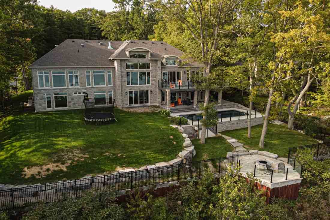 Large stone house with a manicured lawn, stone patio, and wooded lakeside setting