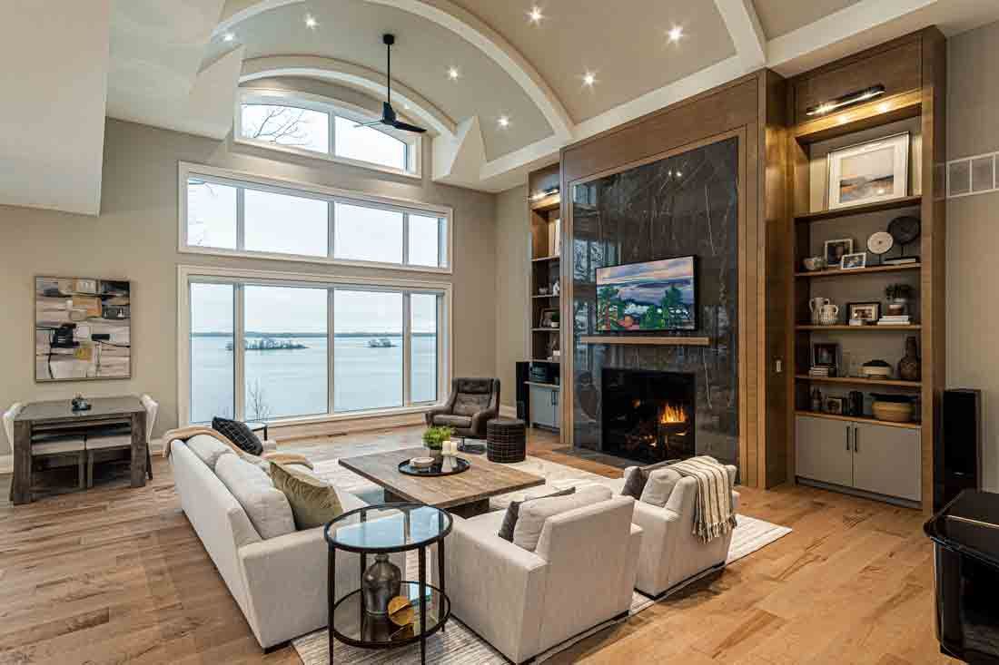 Bright living room with white sectional, stone fireplace, tall windows, and built-in shelves overlooking water.