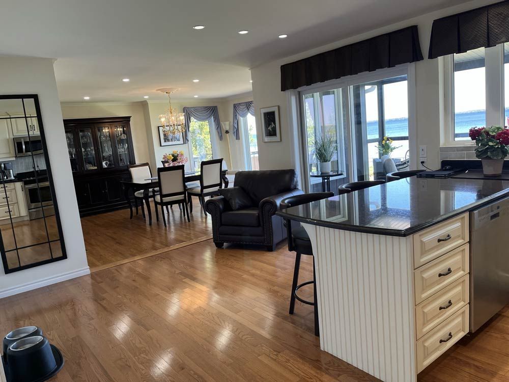 Bright open kitchen and living room with hardwood floors, black chairs, and large windows overlooking water