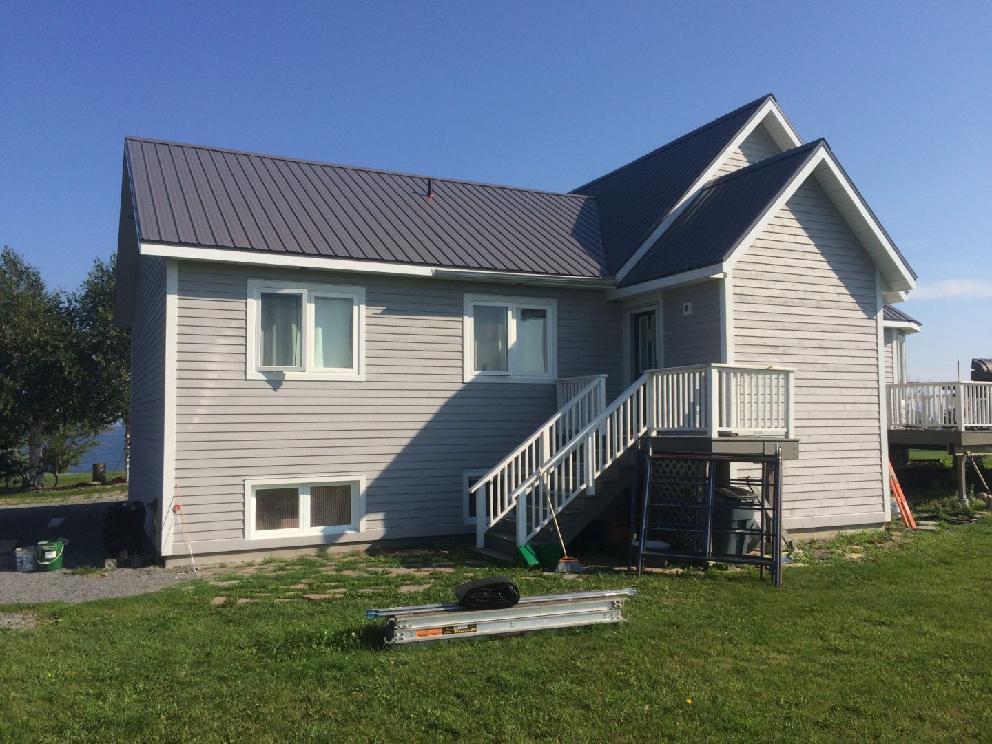 Gray house with metal roof, front porch stairs, and green lawn under a blue sky