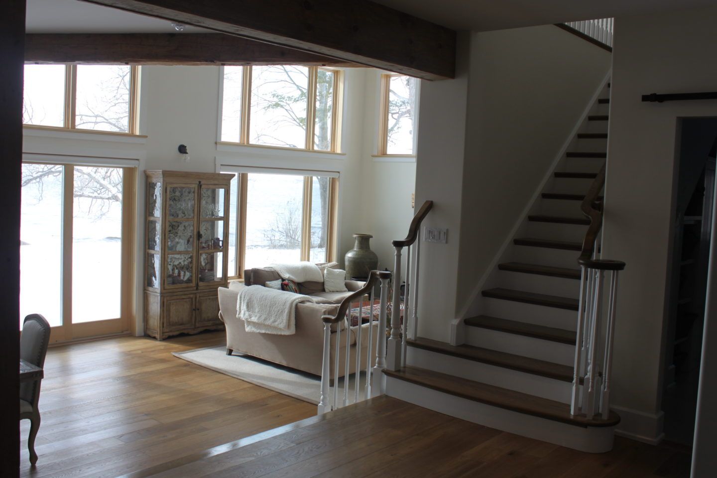Bright living room with large windows, wooden floors, couch, and staircase by the entryway