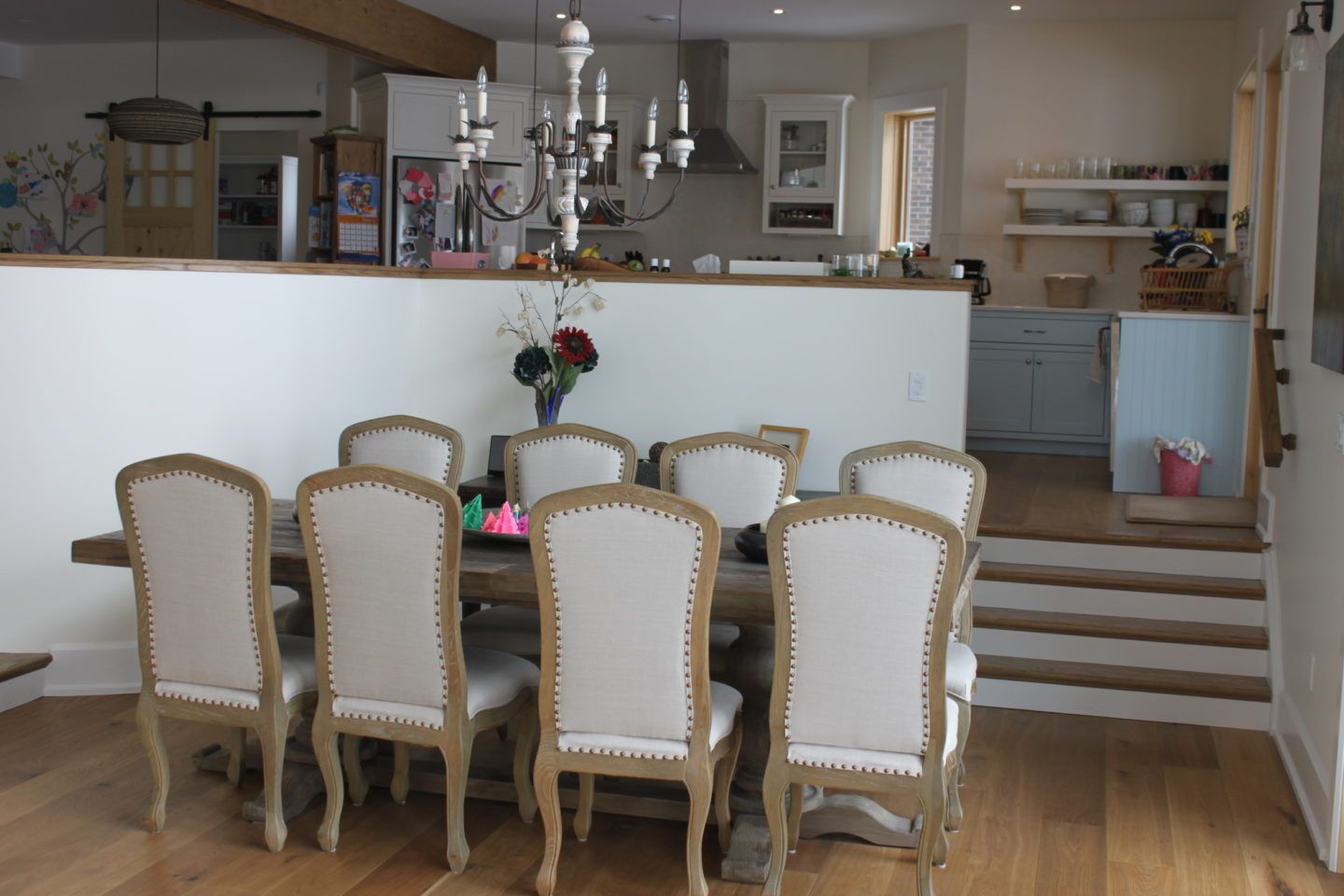 Dining room with six upholstered chairs around a wooden table, facing an open kitchen and white half wall.