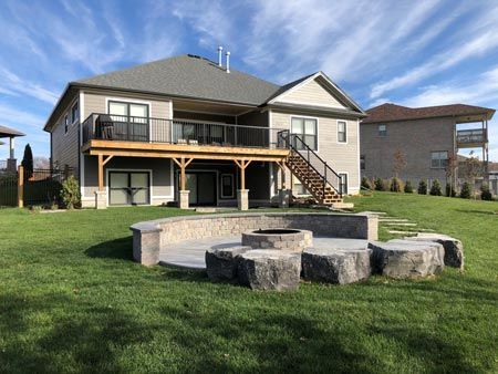 Suburban backyard with stone fire pit, green lawn, and elevated deck on a beige house under a blue sky