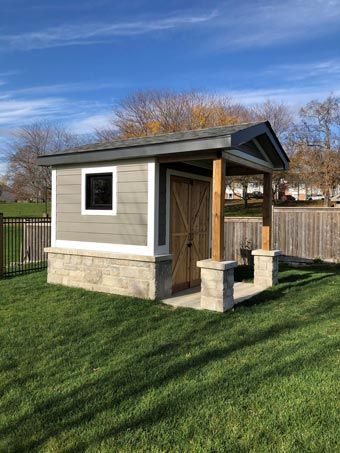 Small gray shed with stone base and wooden door in a grassy backyard under a blue sky