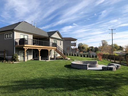 Suburban backyard with deck, stairs, and circular fire pit on a large green lawn.