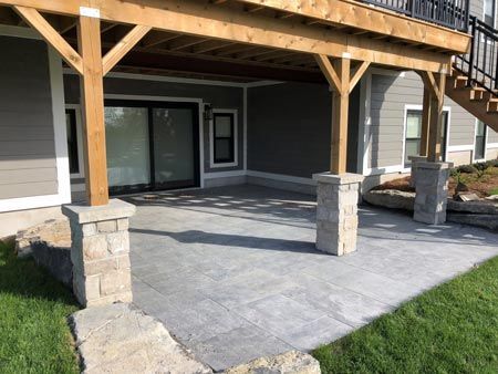 Covered patio with stone columns and gray stamped concrete beside a house