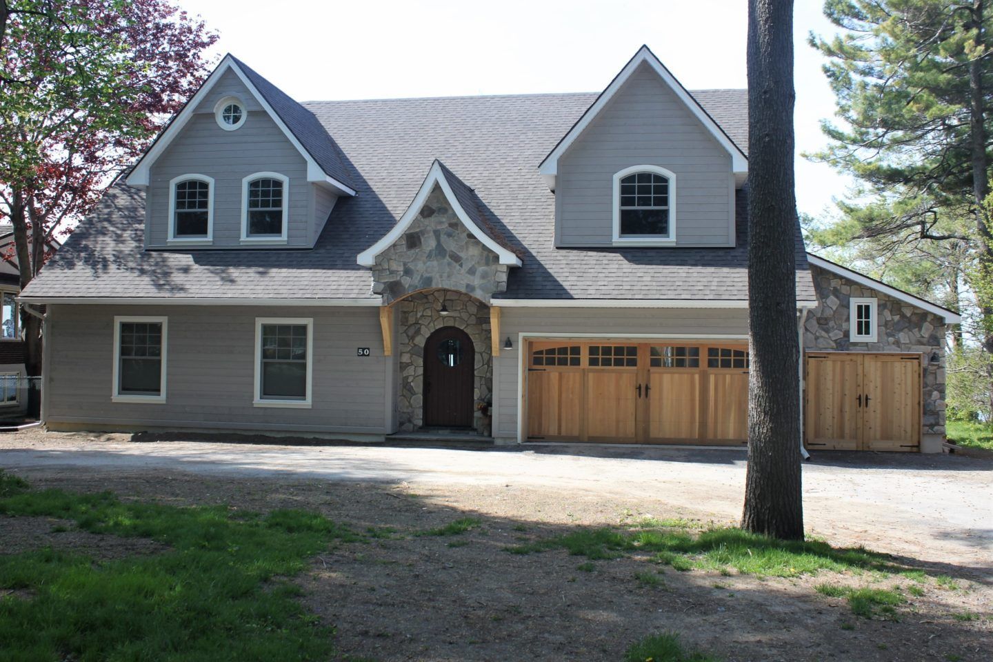 Large gray house with stone accents and a wooden garage door, seen from the front in a wooded yard
