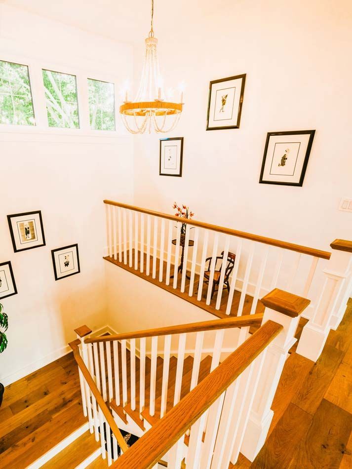 Stairwell with white walls, wood railings and floor, framed art, and a chandelier by a bright window.
