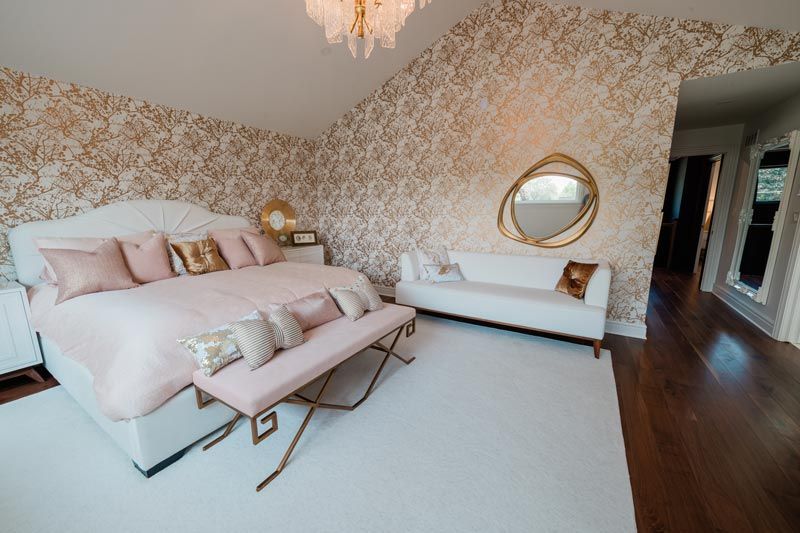 Elegant pink bedroom with floral wallpaper, upholstered bed, bench, and round mirror on a white rug.