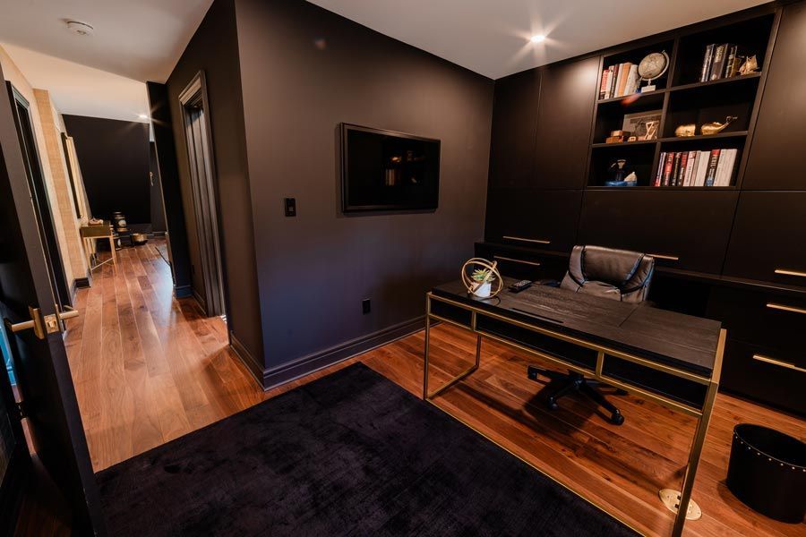Modern home office with dark walls, a desk, shelving, and hardwood floor; hallway visible to the left
