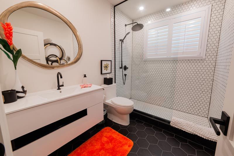 Modern bathroom with white vanity, round mirror, black hex floor, orange rug, and tiled shower with bench