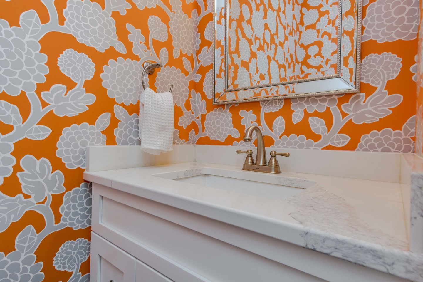 Orange floral wallpaper behind a white marble bathroom vanity with gold faucet and mirror frame