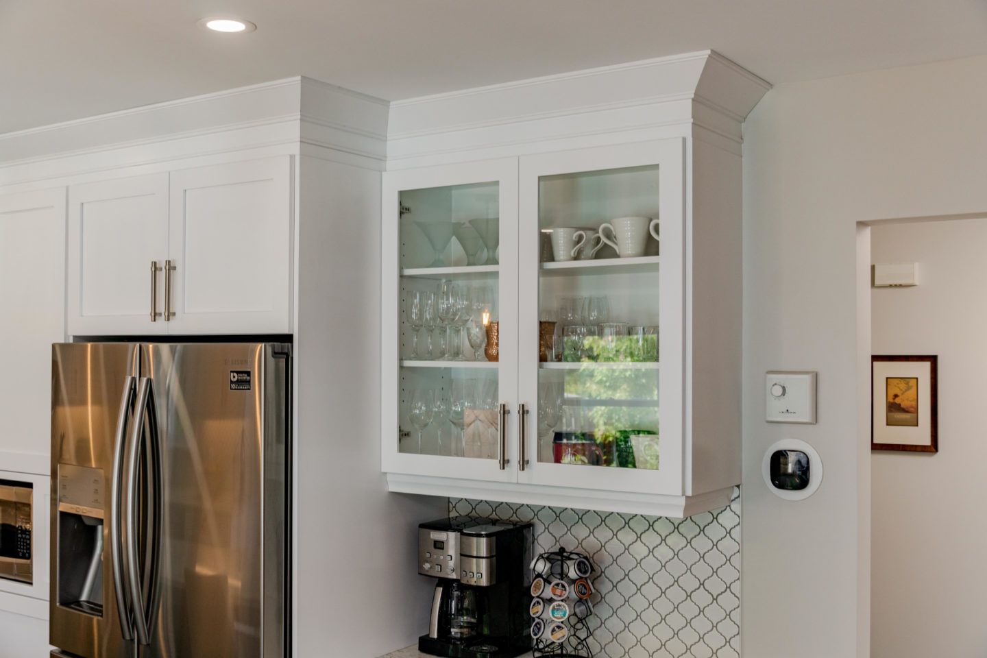 Bright kitchen with white cabinets, glass-front cupboard, stainless fridge, and coffee maker on tiled backsplash.