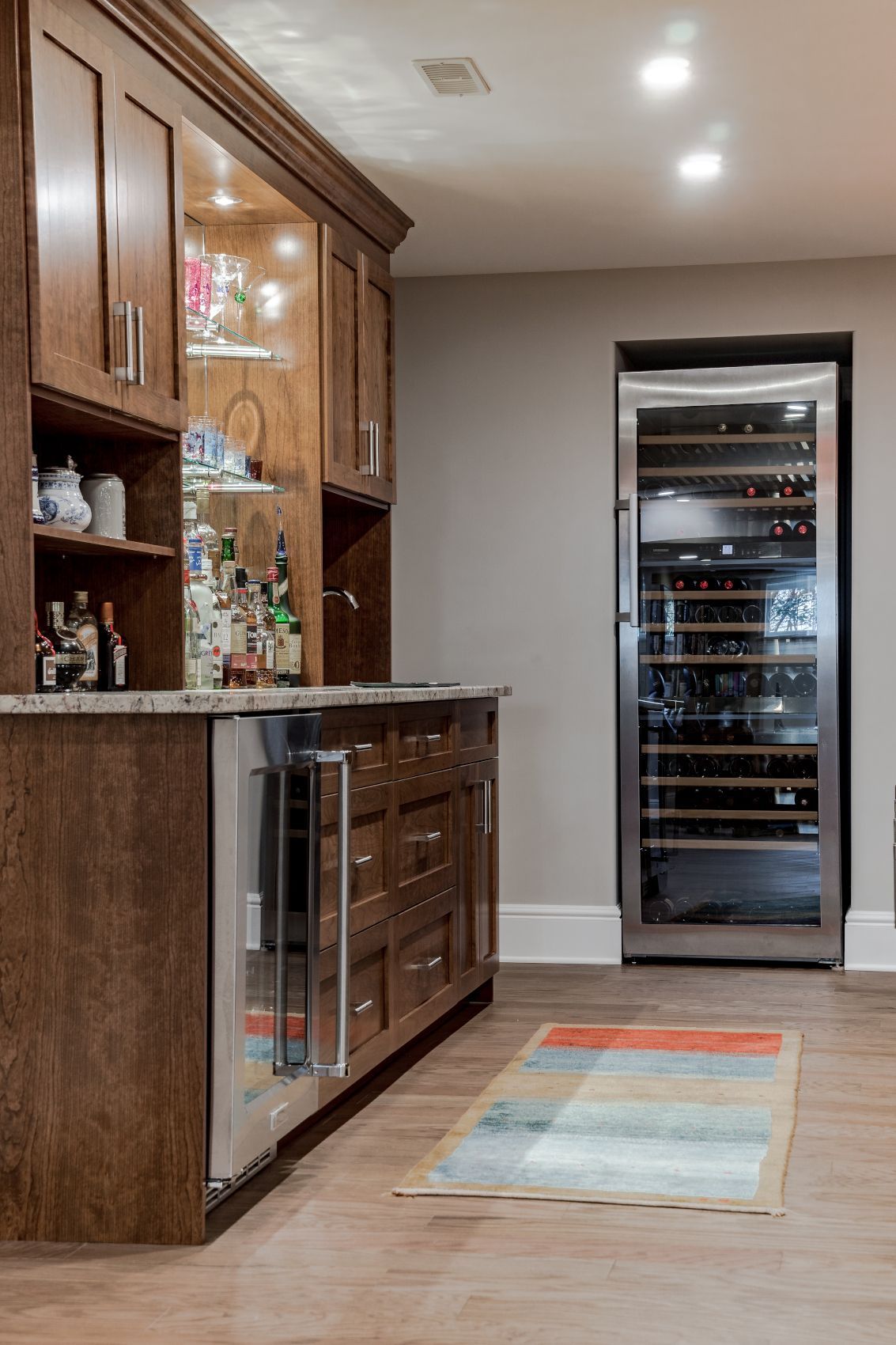 Wood-paneled home bar with a granite counter, wine fridge, and striped rug on a light wood floor.