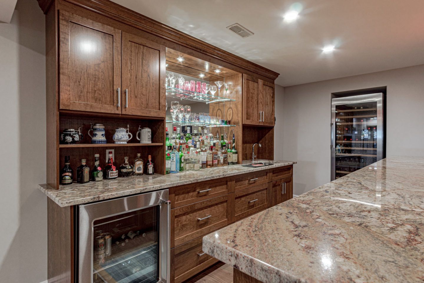 Built-in home bar with wood cabinets, granite countertop, mirror, and glass shelves with bottles and glassware