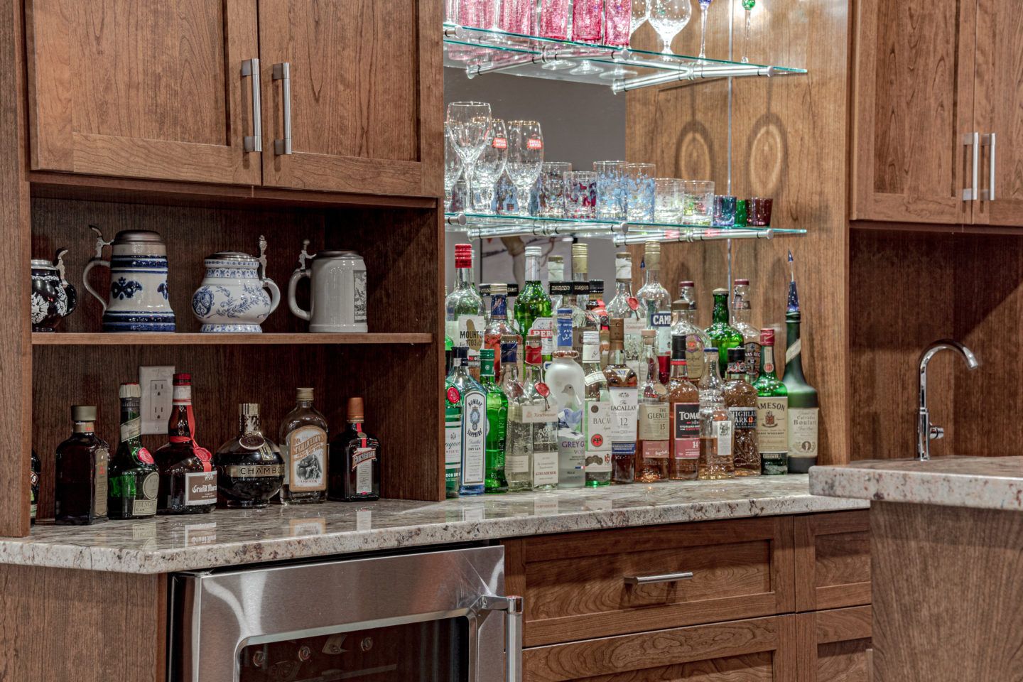 Home bar with wooden cabinets, glass shelves, and assorted bottles and glassware on a granite counter.