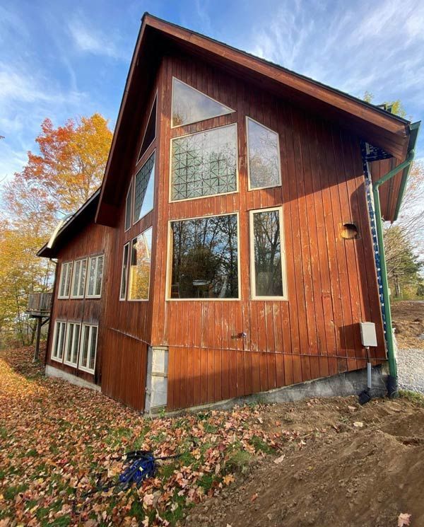 Modern wooden house with large windows and a steep roof, surrounded by autumn trees and fallen leaves.