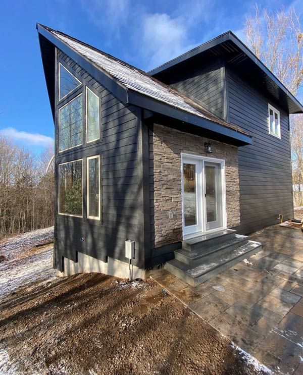 Modern black house exterior with angular roof, large windows, and a small front patio beside bare trees