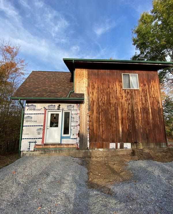 Small rustic building with white and wood siding, a front door, and gravel driveway under a blue sky