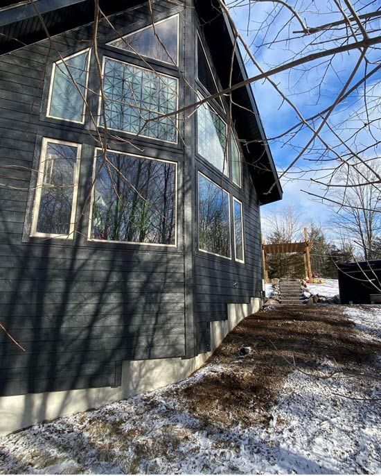 Exterior of a dark gray house with large windows, a snowy ground, and leafless trees on a sunny day