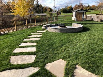 Backyard with stepping stones leading to a circular fire pit and small shed, bordered by a wooden fence and trees
