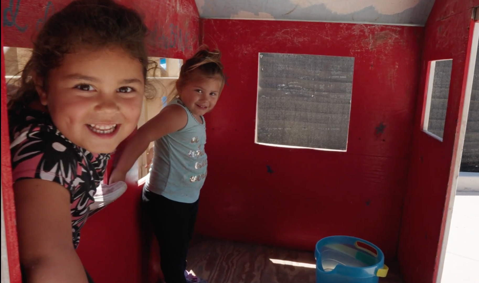 Two children smiling inside a small red playhouse with a window and blue bucket