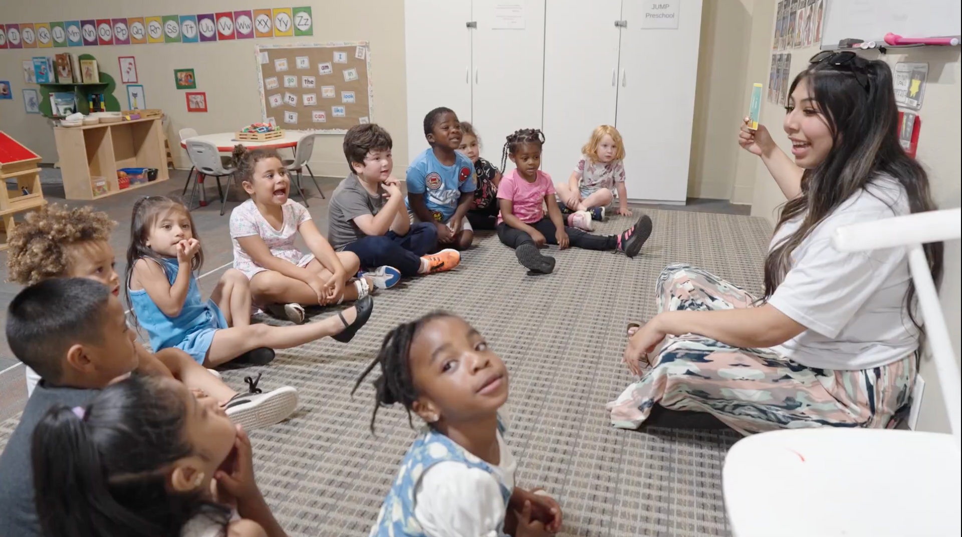 Teacher reading to a circle of children sitting on a classroom rug, with shelves and toys in the background