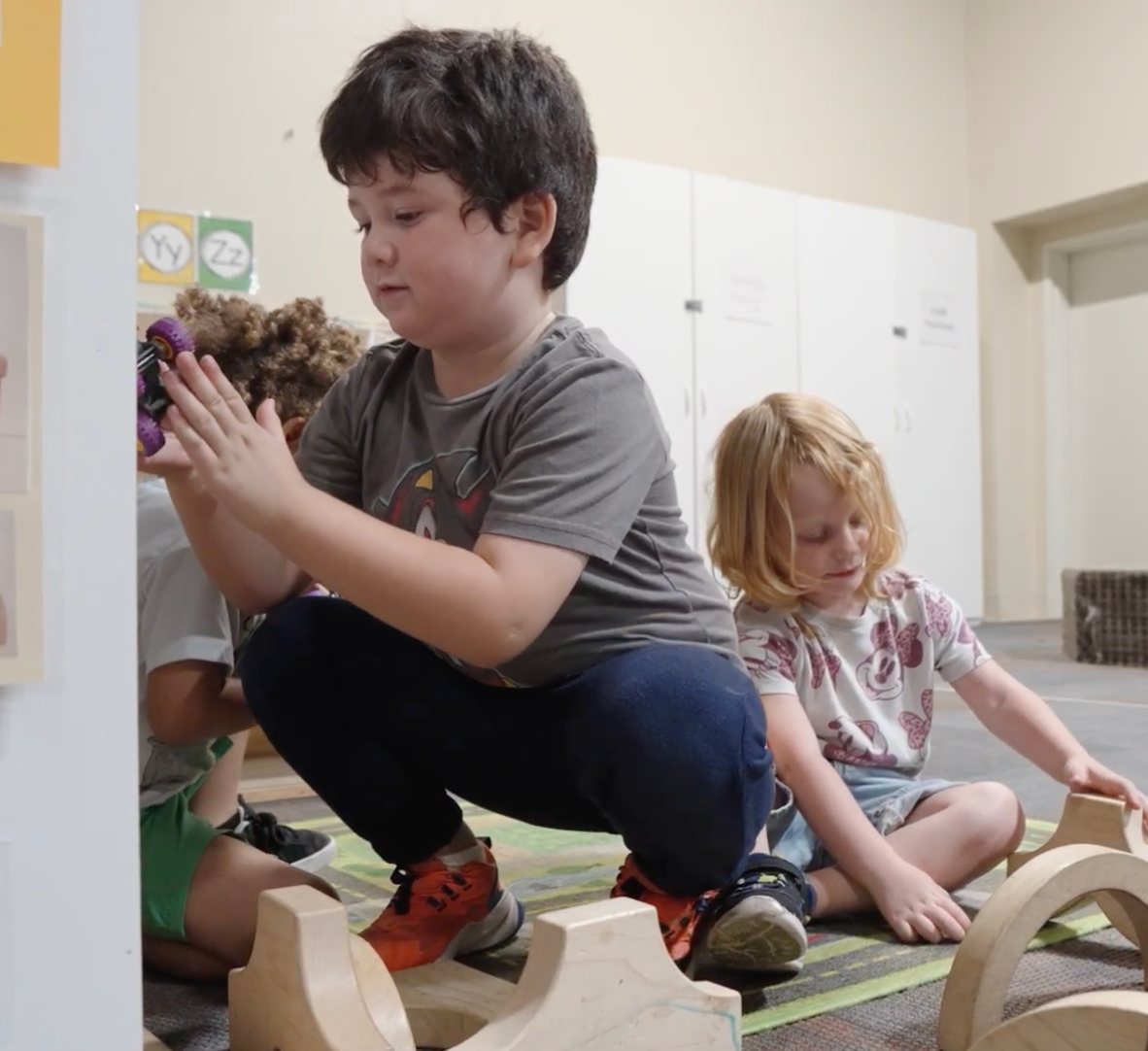 Children playing with toys on the floor in a bright classroom, one crouching and another seated nearby
