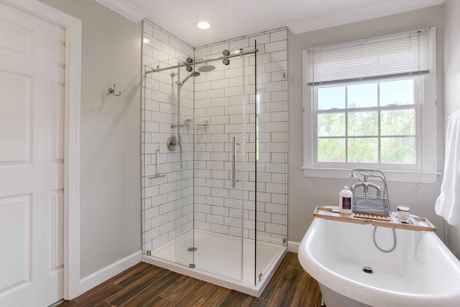 Bathroom with glass shower, white tub, window, and hardwood floor.