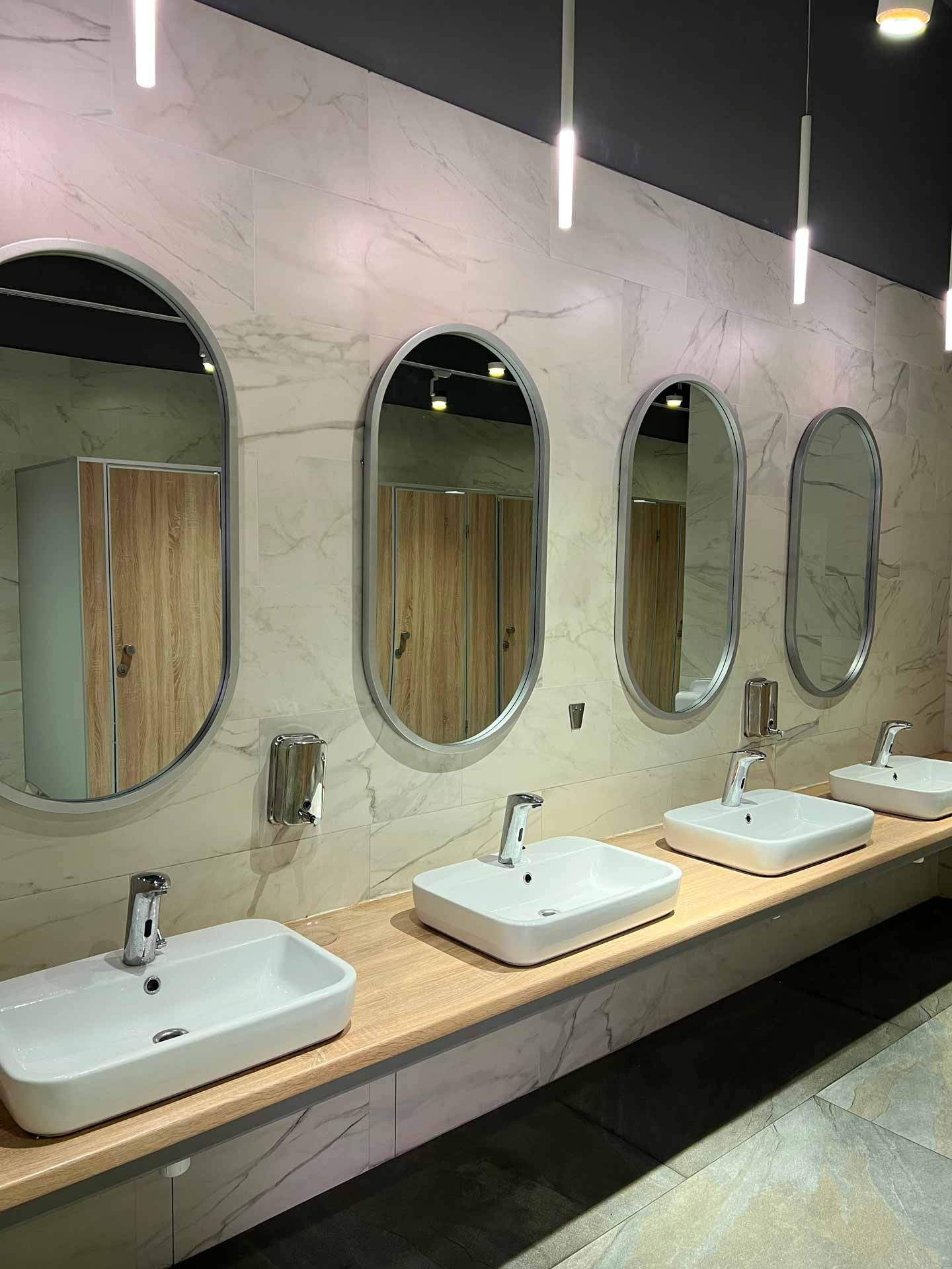 Public restroom with white sinks, oval mirrors, and wood stalls in Teaneck, NJ