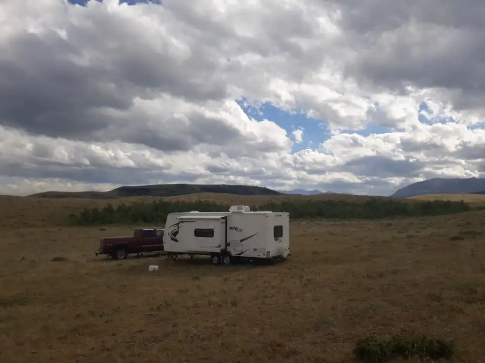 White travel trailer and truck parked in a grassy field under a cloudy sky with distant hills.