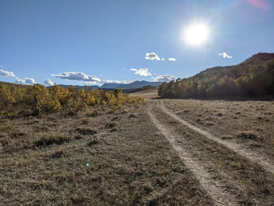 Sunny dirt trail through a dry grassy field with hills and mountains in the distance