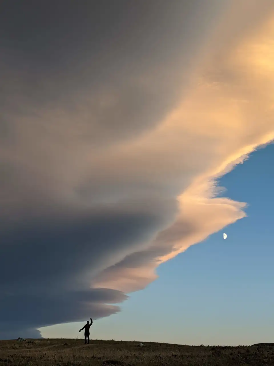 Massive layered storm cloud over a lone person standing on a hill at sunset