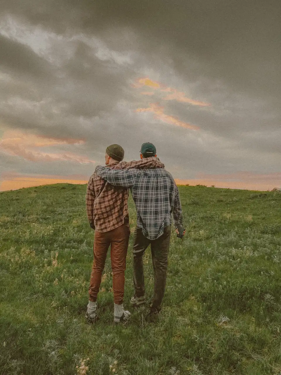 Two people standing arm-in-arm in a grassy field under a cloudy sunset sky