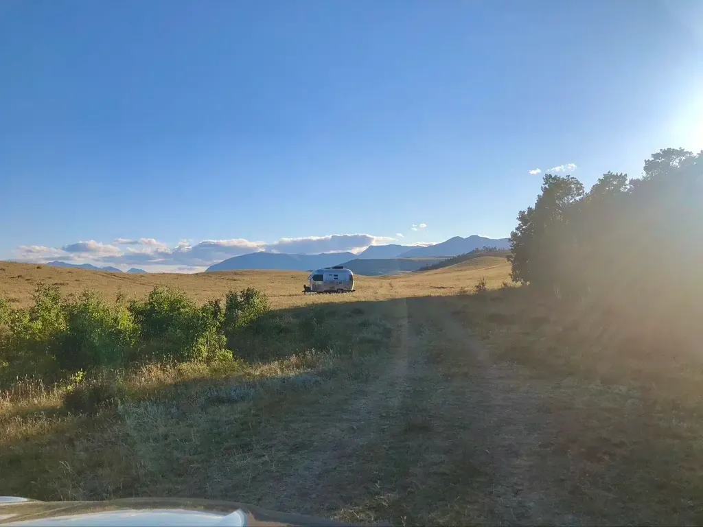 White van on a dirt track in a grassy field, with mountains under a clear blue sky and bright sun glare.