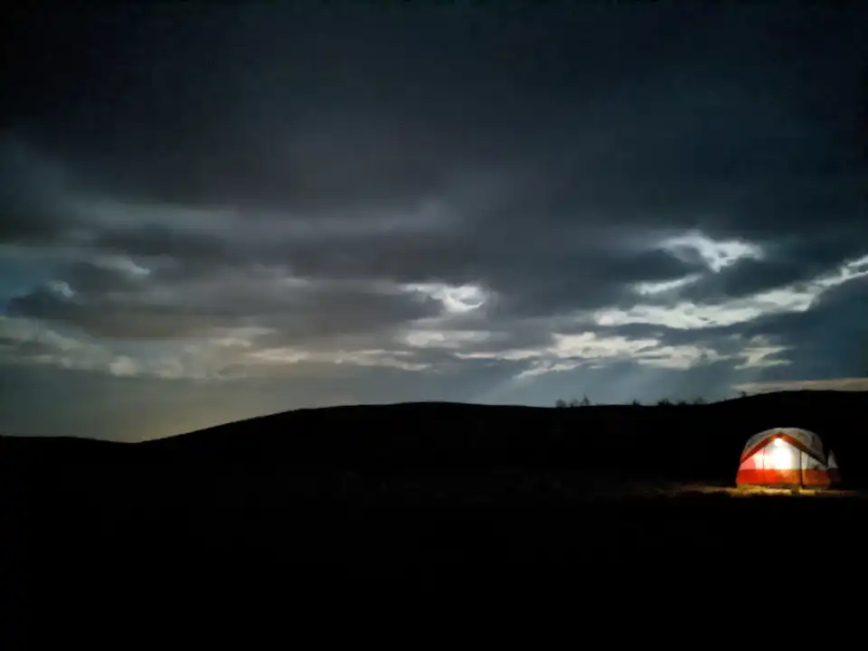 Dark night sky over a silhouetted hill, with a glowing orange tent at the lower right.